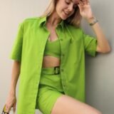 Комплект женской льняной одежды "Alessandra" салатового цвета, состоящий из рубашки с коротким рукавом, топа, шорт и пояса, на девушке, сидящей в кресле. Комплект жіночого лляного одягу "Alessandra" салатового кольору, що складається з сорочки з коротким рукавом, топа, шортів і пояса, на дівчині, яка сидить у кріслі.